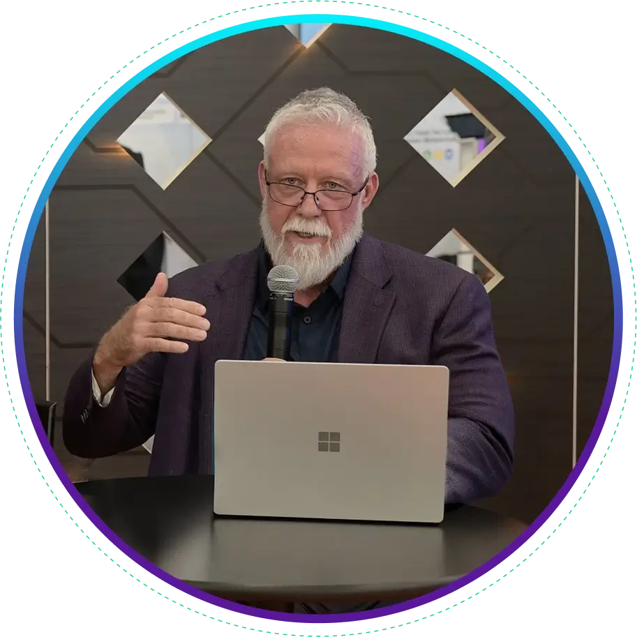 A middle-aged man with a beard and glasses speaking into a microphone, gesturing with his hand, with a Microsoft laptop on the table in front of him.